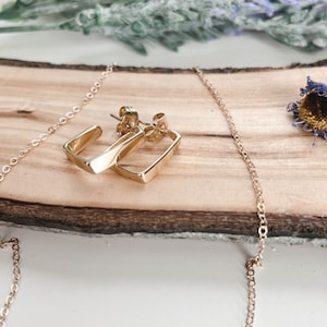 May include: A gold chain necklace and a pair of gold square hoop earrings on a wooden surface.