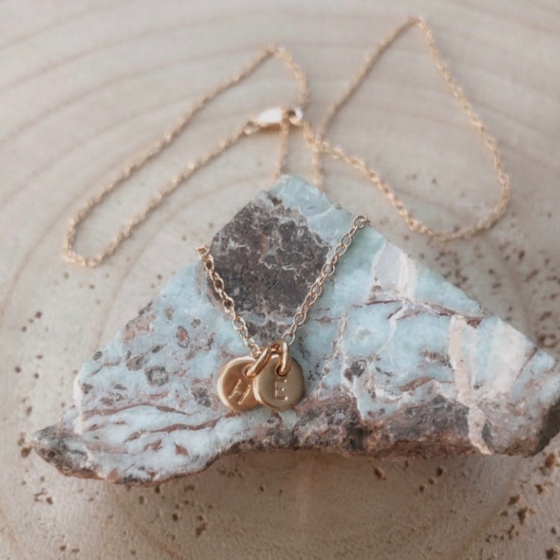 May include: A gold chain necklace with two small gold discs, each with a letter engraved on them. The letters are "H" and "E".