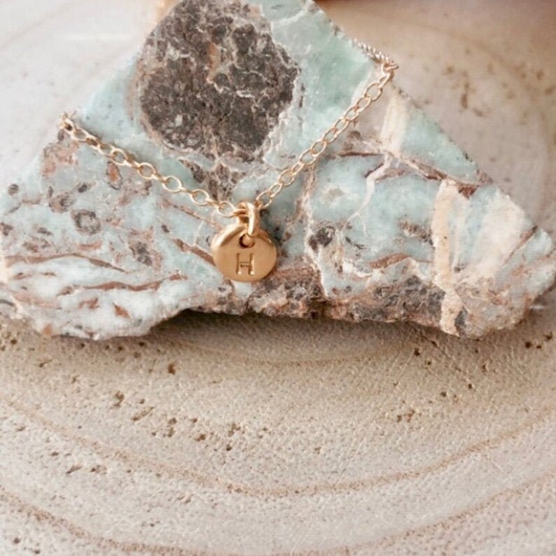 May include: A gold chain necklace with a small gold charm featuring the letter "H". The necklace is displayed on a light brown and green rock.