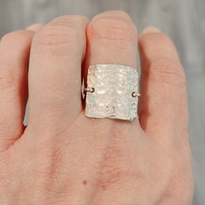 May include: A silver square ring with a hammered texture and the word "BRODIE" engraved on the front.