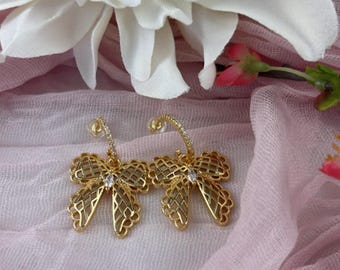 May include: A pair of gold butterfly-shaped hoop earrings with clear crystal accents. The earrings are on a pink fabric background.