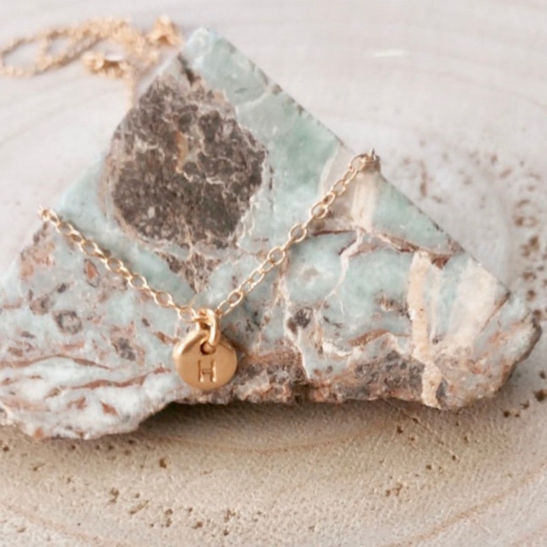 May include: A delicate gold chain necklace with a small, round pendant featuring the letter "H" engraved on it. The necklace is displayed on a light green and brown stone.