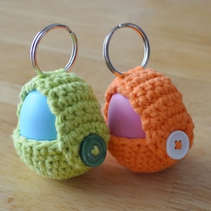 Könnte beinhalten: Zwei handgefertigte Schlüsselanhänger mit gehäkelten Hüllen in Grün und Orange, jeweils mit Knopfverschluss. Der grüne Schlüsselanhänger enthält ein blaues Objekt, der orangefarbene ein rosa Objekt. Beide haben silberne Schlüsselringe.