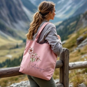 May include: A pink tote bag with a floral design and the name "Abigail" embroidered on it. The bag has long straps and is made of a soft material. The floral design features light pink and green flowers.