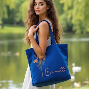 May include: A cobalt blue tote bag featuring a floral illustration and the name "Emma" in a cursive script. The bag has strong handles and is made of a robust material. A heart symbol is next to the name.