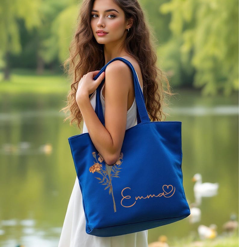 May include: A cobalt blue tote bag featuring a floral illustration and the name "Emma" in a cursive script. The bag has strong handles and is made of a robust material. A heart symbol is next to the name.
