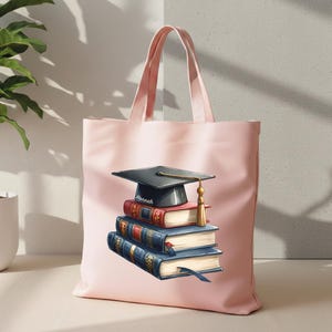 May include: A light pink tote bag featuring a stack of books topped with a graduation cap. The books are red and blue with gold accents. The graduation cap is black with a gold tassel. The name "Hannah" is on the cap.