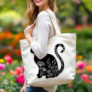 K&ouml;nnte beinhalten: Eine wei&szlig;e Einkaufstasche mit einer schwarzen Silhouette einer Katze mit floralen Details am Schwanz. Die Katze sitzt mit ihrem Schwanz um ihren K&ouml;rper gewickelt.