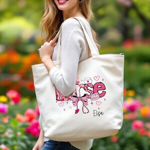May include: Cream-colored tote bag with the word "Nurse" in pink and white, decorated with hearts, a stethoscope, and a bow. The bag has a name, "Elsa," printed in black. The bag is carried by a person.