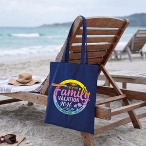 May include: A navy blue tote bag with the text "Choplin Family Vacation 2026" in a colorful design. The bag is hanging on a wooden beach chair, with a straw hat and sunglasses nearby. The background shows the ocean and sand.