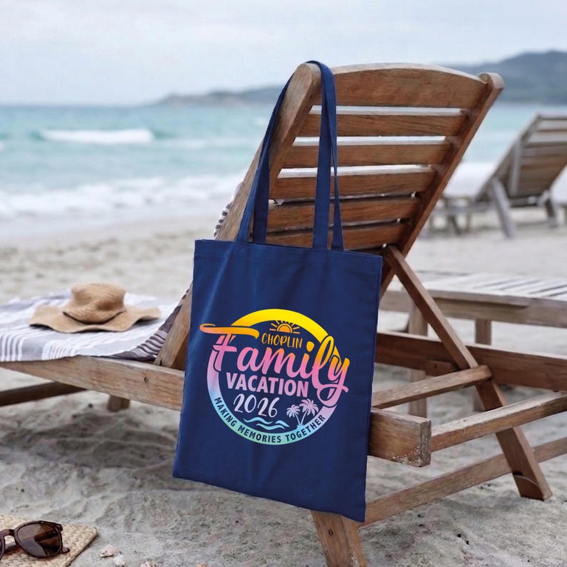 May include: A navy blue tote bag with the text "Choplin Family Vacation 2026" in a colorful design. The bag is hanging on a wooden beach chair, with a straw hat and sunglasses nearby. The background shows the ocean and sand.