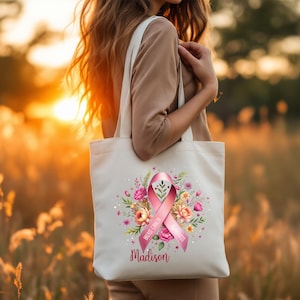 May include: Cream-colored tote bag featuring a pink ribbon with the words "Breast Cancer Awareness" and a floral design. The name "Madison" is written in cursive below the ribbon. The bag has cream-colored handles.