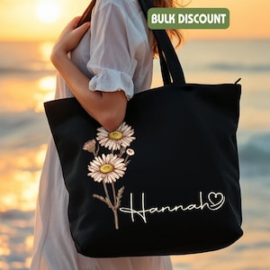 May include: Black tote bag with a daisy floral design and the name "Hannah" in white script. The bag has long handles. The background is a blurred beach scene. The words "Bulk Discount" are in a green rectangle.
