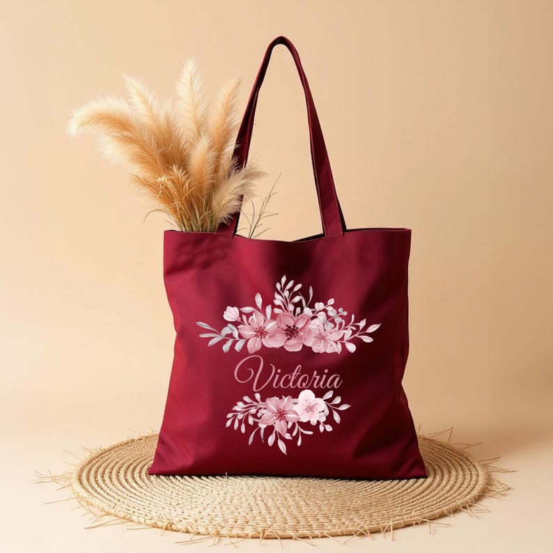 May include: A burgundy tote bag with a floral design and the name "Victoria" printed in pink. The bag is made of canvas and has two handles.