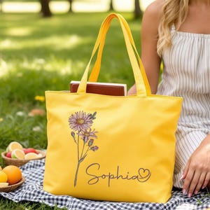 May include: A vibrant yellow tote bag with a floral design and the name "Sophia" in script. The bag, filled with books, is set on a checkered blanket. The scene suggests a picnic or outdoor leisure activity.