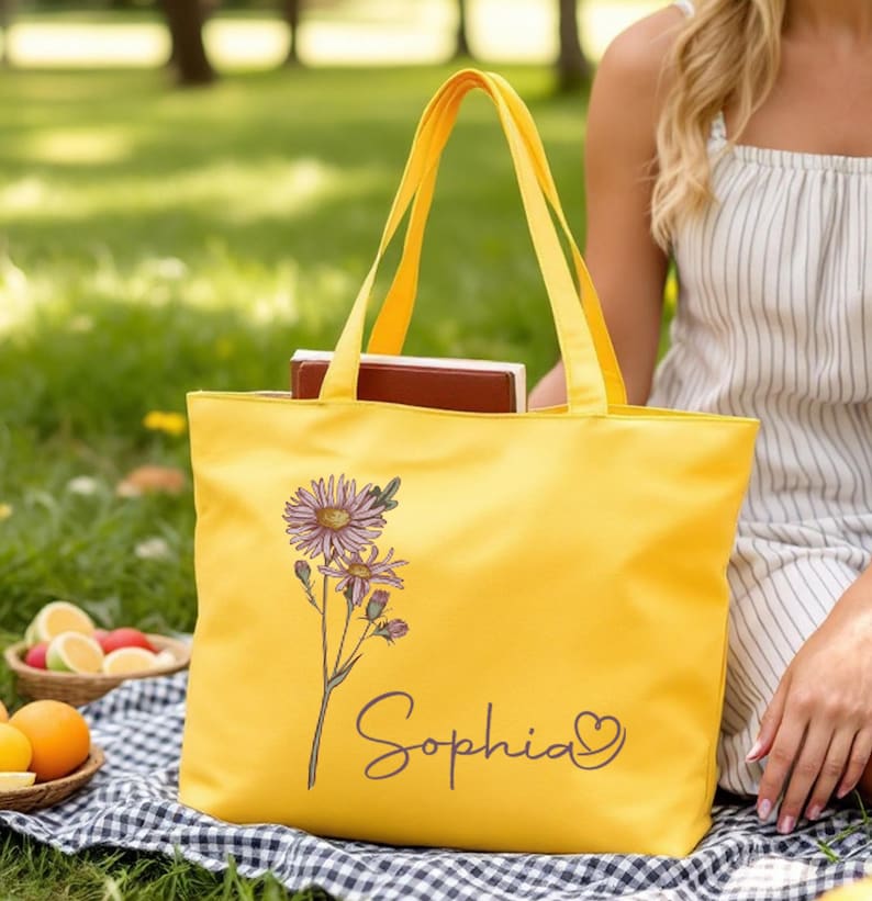 May include: A vibrant yellow tote bag with a floral design and the name "Sophia" in script. The bag, filled with books, is set on a checkered blanket. The scene suggests a picnic or outdoor leisure activity.