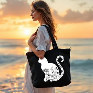 K&ouml;nnte beinhalten: Eine schwarze Tragetasche mit einer wei&szlig;en Silhouette einer Katze mit floralen Details. Die Tasche hat einen Rei&szlig;verschluss.
