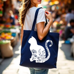 K&ouml;nnte beinhalten: Eine marineblaue Tragetasche mit einer wei&szlig;en Silhouette einer Katze mit floralen Details. Der Schwanz der Katze ist um die Blumen gewickelt.