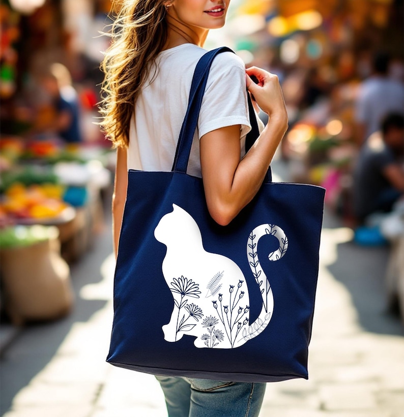 K&ouml;nnte beinhalten: Eine marineblaue Tragetasche mit einer wei&szlig;en Silhouette einer Katze mit floralen Details. Der Schwanz der Katze ist um die Blumen gewickelt.