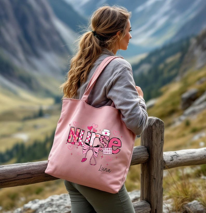 May include: A pink tote bag with the word "Nurse" in a decorative font, featuring hearts, a bow, and a stethoscope. The bag has long straps and the name "Luna" is printed below the word "Nurse".