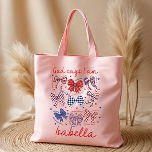 May include: A light pink tote bag with a design featuring bows and the text "God says I am." The bows are in red, white, and blue. The name "Isabella" is written in red script at the bottom. The bag has two handles.