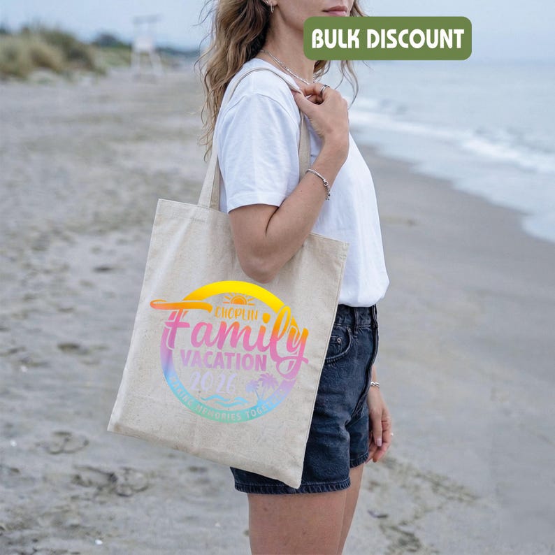 May include: A beige canvas tote bag with a colorful graphic that reads "European Family Vacation 2026". The design features a sunset, palm trees, and waves. The bag is being held by a person on a beach.