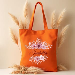 May include: Orange canvas tote bag with a floral design and the name "Isabella" printed in black.