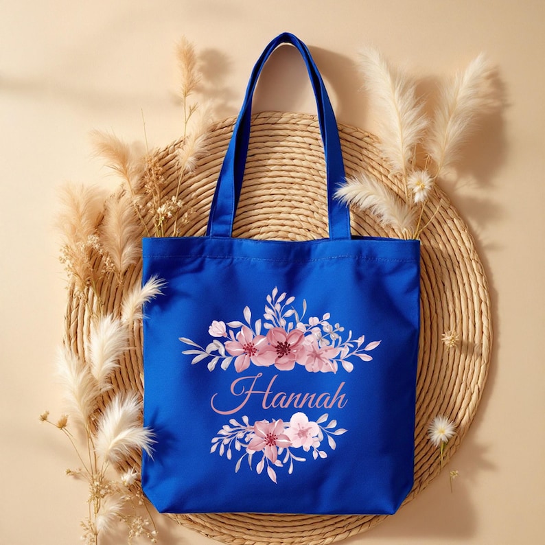 May include: A royal blue tote bag with a floral design and the name "Hannah" printed in pink. The bag is sitting on a woven straw mat.