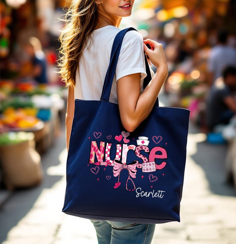 May include: A navy blue tote bag with the word "NURSE" in pink and patterned letters, decorated with hearts, a stethoscope, and a nurse's hat. The name "Scarlett" is printed below the design.