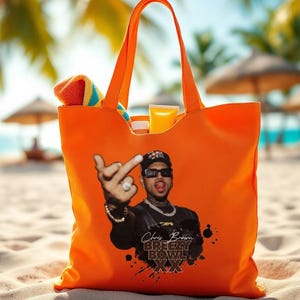 May include: Bright orange tote bag with a graphic of a person wearing sunglasses and a black hat. The bag has the text "Chris Brown Breezy Bowl XX". The bag is filled with a towel and sunscreen, perfect for a day at the beach.