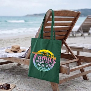 May include: A dark green tote bag with the words "Choplin Family Vacation 2026" in a colorful design. The bag is on a wooden beach chair with a straw hat and sunglasses nearby. The beach and ocean are in the background.