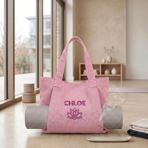 May include: A pink canvas tote bag featuring the name CHLOE and a lotus flower graphic printed in a darker shade of pink. The bag has two shoulder straps and a grey yoga mat secured horizontally through the center of the bag's exterior.