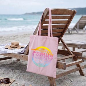 May include: A light pink tote bag with long handles, featuring a colorful circular design that reads "Family Vacation 2026". The bag is made of a soft fabric and is sitting on a wooden beach chair near the ocean.