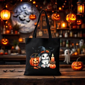 May include: Black tote bag featuring a Halloween-themed design. The graphic includes a white bunny wearing a witch hat, sitting next to a jack-o'-lantern, candle, and pumpkins. Bats and a carved pumpkin are in the background.