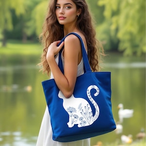 K&ouml;nnte beinhalten: Eine blaue Tragetasche mit einer wei&szlig;en Silhouette einer Katze mit floralen Details. Der Schwanz der Katze ist um eine Blume gewickelt.