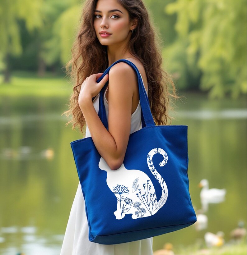 K&ouml;nnte beinhalten: Eine blaue Tragetasche mit einer wei&szlig;en Silhouette einer Katze mit floralen Details. Der Schwanz der Katze ist um eine Blume gewickelt.