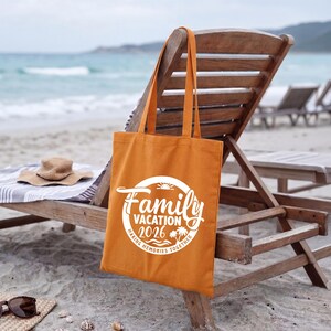 May include: An orange tote bag with the words "Family Vacation 2026" in a white circular design. The bag is hanging on a wooden beach chair. A straw hat and sunglasses rest on the sand.