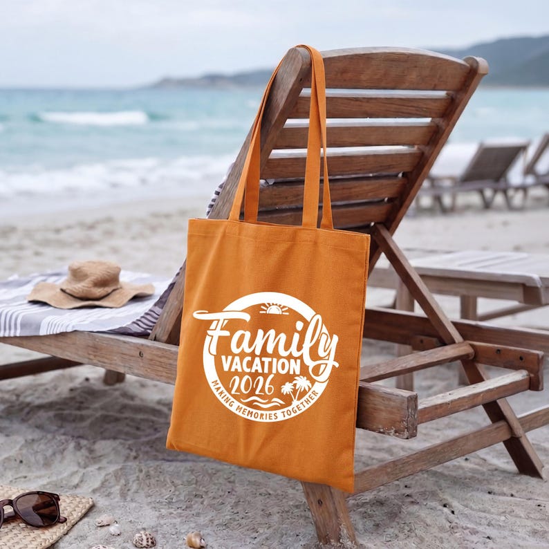 May include: An orange tote bag with the words "Family Vacation 2026" in a white circular design. The bag is hanging on a wooden beach chair. A straw hat and sunglasses rest on the sand.