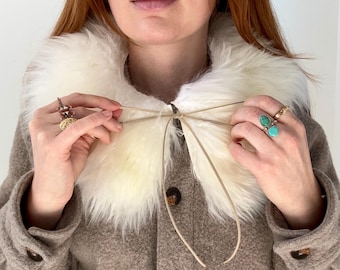 May include: A detachable white faux fur collar with thin beige tie strings, worn over a tan button-down jacket. The collar features long, soft synthetic fibers and is secured at the center front with a simple bow tie closure.