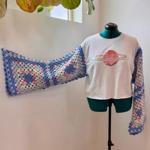 May include: A white crop top with a pink planet graphic and crocheted sleeves. The sleeves are made of blue, pink, and white granny squares. The top is displayed on a green dress form.