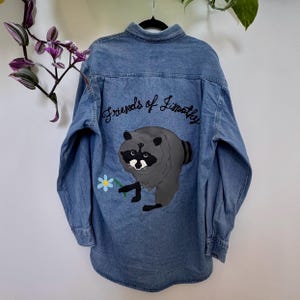 May include: A light blue denim button-down shirt featuring a graphic of a gray raccoon holding a light blue flower with a yellow center. Above the raccoon, the text Friends of Timothy is embroidered in black cursive script on the back panel.