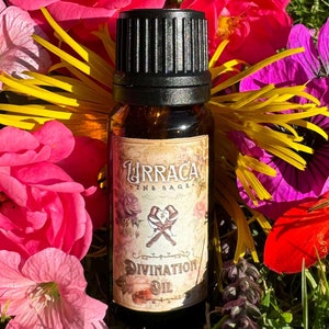 Op de afbeelding: Een klein bruin glazen flesje met een zwarte dop en een etiket met de tekst "Turraca The Sage" en "Divination Oil". Het flesje is omringd door roze, gele, paarse en rode bloemen.