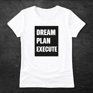 May include: White t-shirt with the words "DREAM PLAN EXECUTE" in bold, white capital letters inside a black rectangle. The shirt has a crew neck and short sleeves. The background is a dark gray.