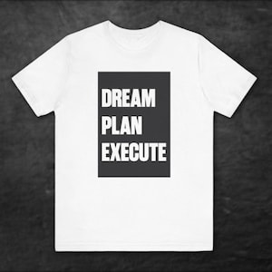 May include: White t-shirt with a black rectangular graphic. The graphic features the words "DREAM PLAN EXECUTE" in bold, white capital letters. The shirt is made of a soft material and has short sleeves.