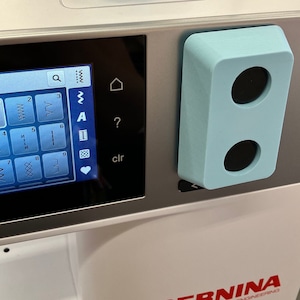 May include: A close-up of a white sewing machine with a blue control panel and a digital display showing stitch options. The display shows numbers 1 through 9 and the letters A and B. The text "clr" is visible on the display.