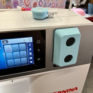 May include: A white sewing machine with a blue and white control panel. The machine has a digital display with a variety of stitch options. The text "BERNINA SWISS ENGINEERING" is printed on the front of the machine.