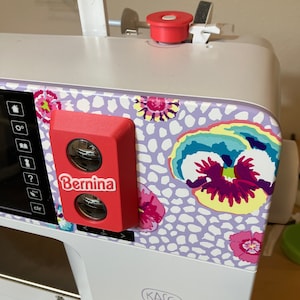 May include: A white sewing machine with a red Bernina logo and a floral design on the side. The machine has a digital display and a variety of buttons and controls. The text "Kaffee Edition B 770 QE Plus" is visible on the front of the machine.
