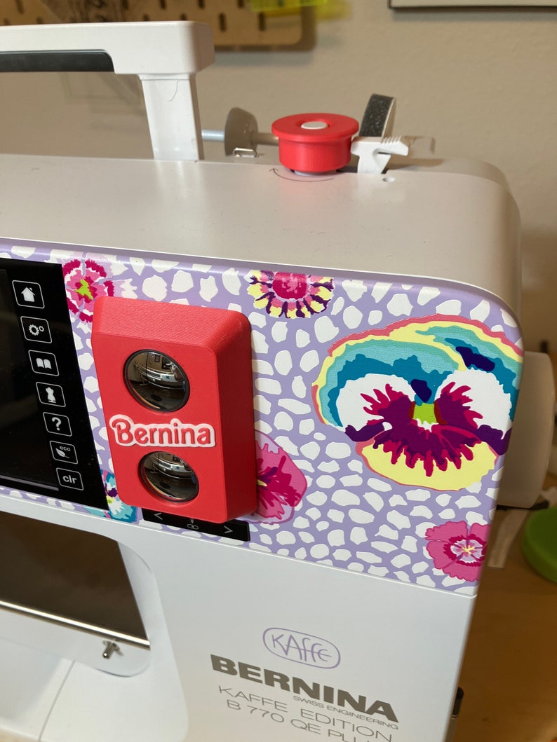 May include: A white sewing machine with a red Bernina logo and a floral design on the side. The machine has a digital display and a variety of buttons and controls. The text "Kaffee Edition B 770 QE Plus" is visible on the front of the machine.