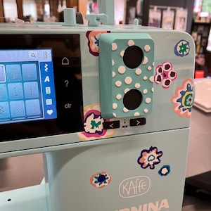 May include: A light blue sewing machine with a white and black polka dot design on the side. The machine has a digital display and a variety of buttons and controls. The text "KAFFE EDITION B 475 QE" is printed on the front of the machine.