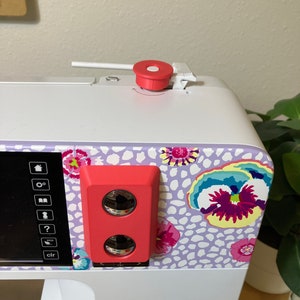 May include: A white sewing machine with a red knob and a red sensor on the side. The machine has a digital display and a floral design on the front. The text "BERNINA Kaffe Edition Swiss Engineering B 770 QE PLUS" is printed on the front of the machine.